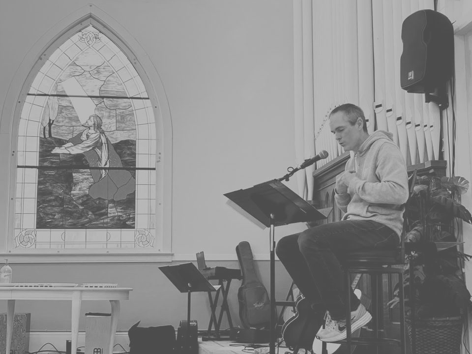 A worship leader singing and playing guitar beside the chapel's stained glass window